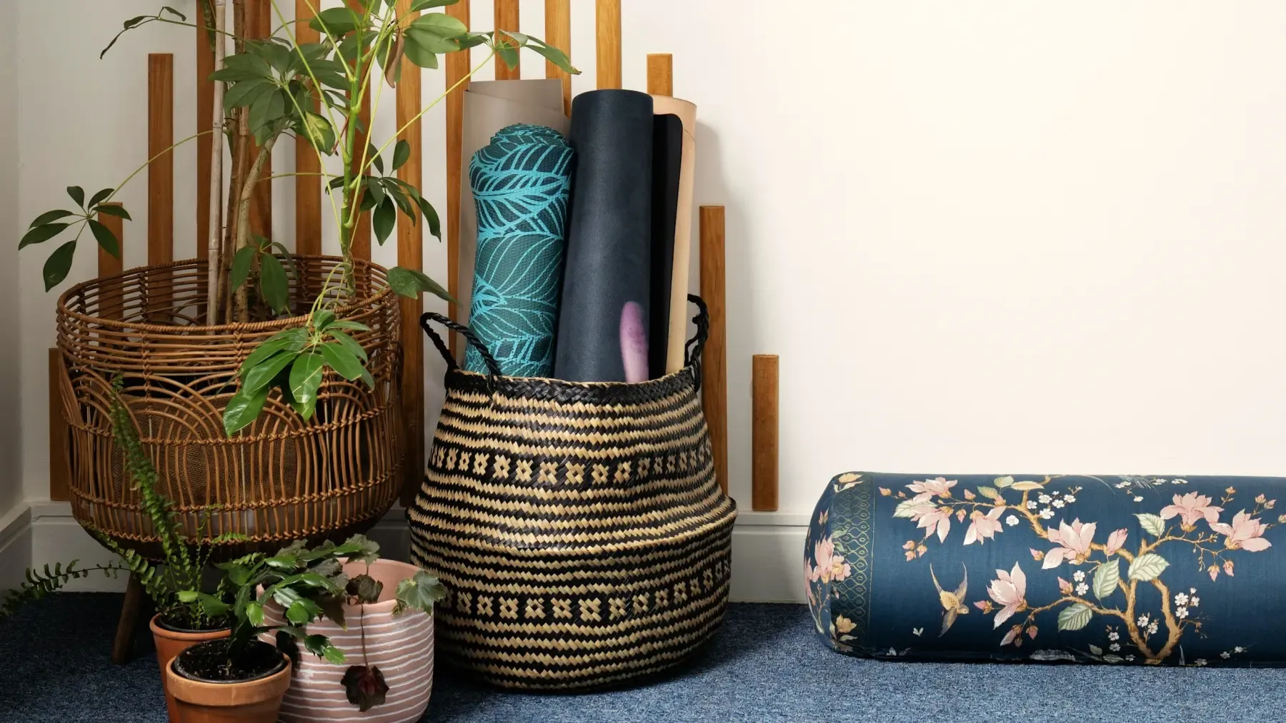 Rolled yoga mats in a woven basket beside plants and a bolster cushion